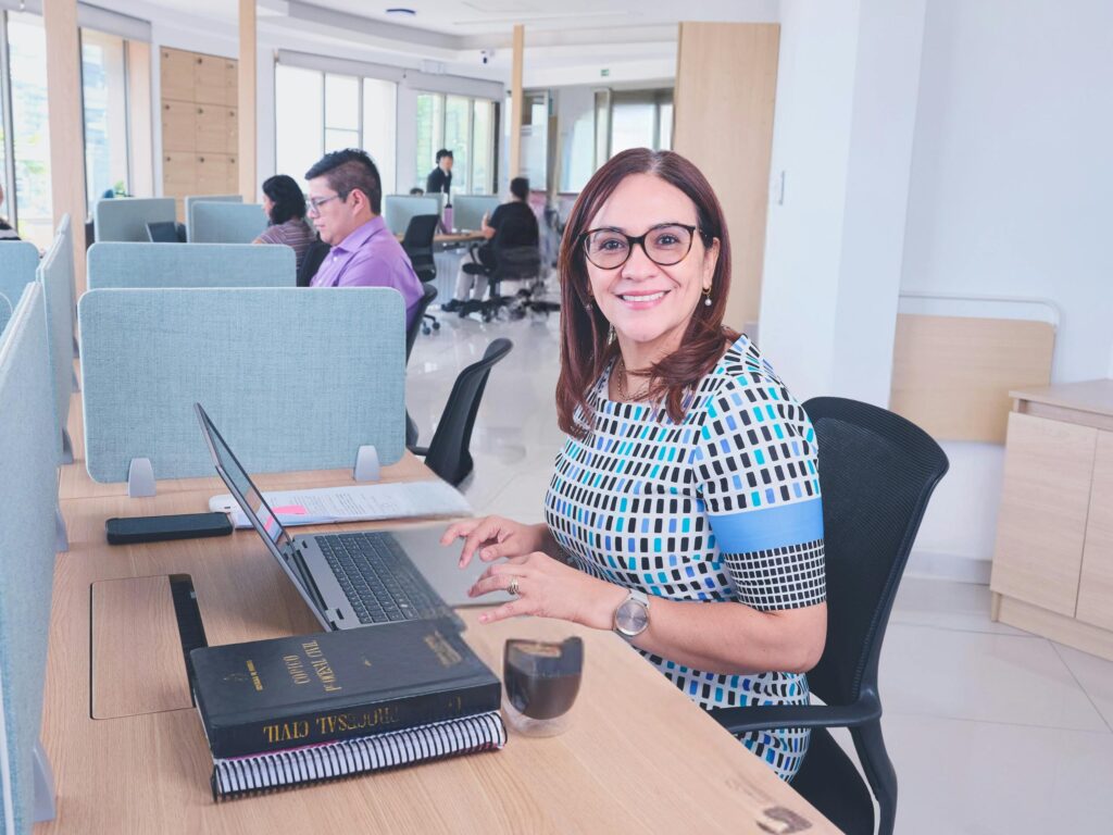 Confident woman smiling while working at an office desk with colleagues nearby.