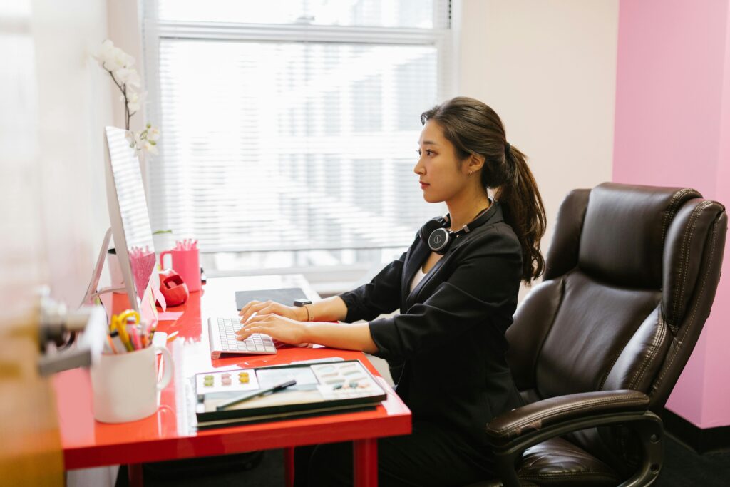 Professional Asian woman engaging in remote work at a stylish office desk with a computer and headphones.